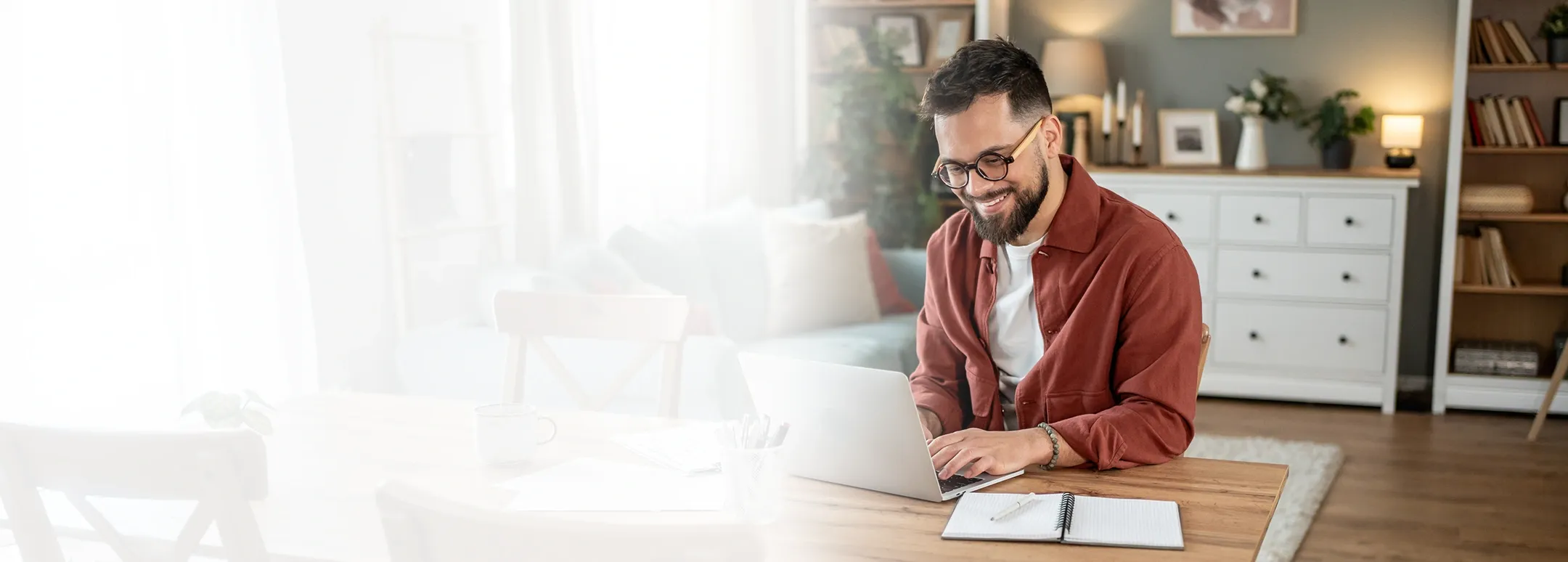 A smiling man with a beard and glasses works on a laptop at a wooden dining table in a bright, modern living space